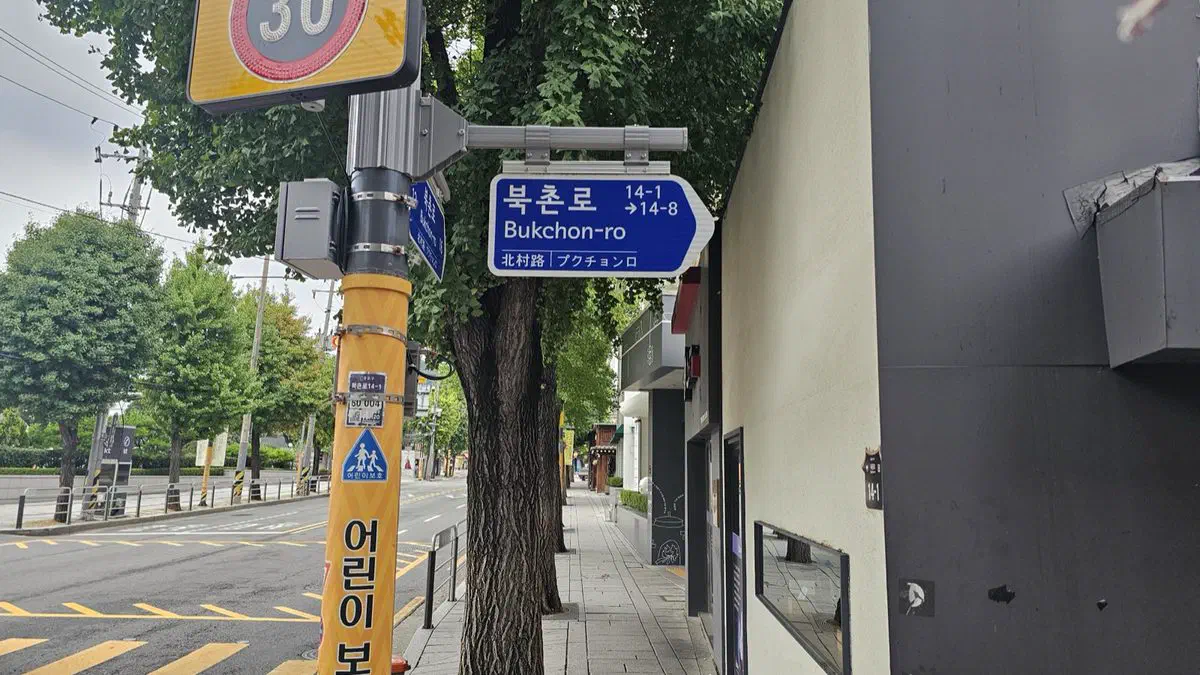Bukchon-ro street sign in both Korean and English mounted above a wide, multi-lane road with a marked crosswalk and passing traffic