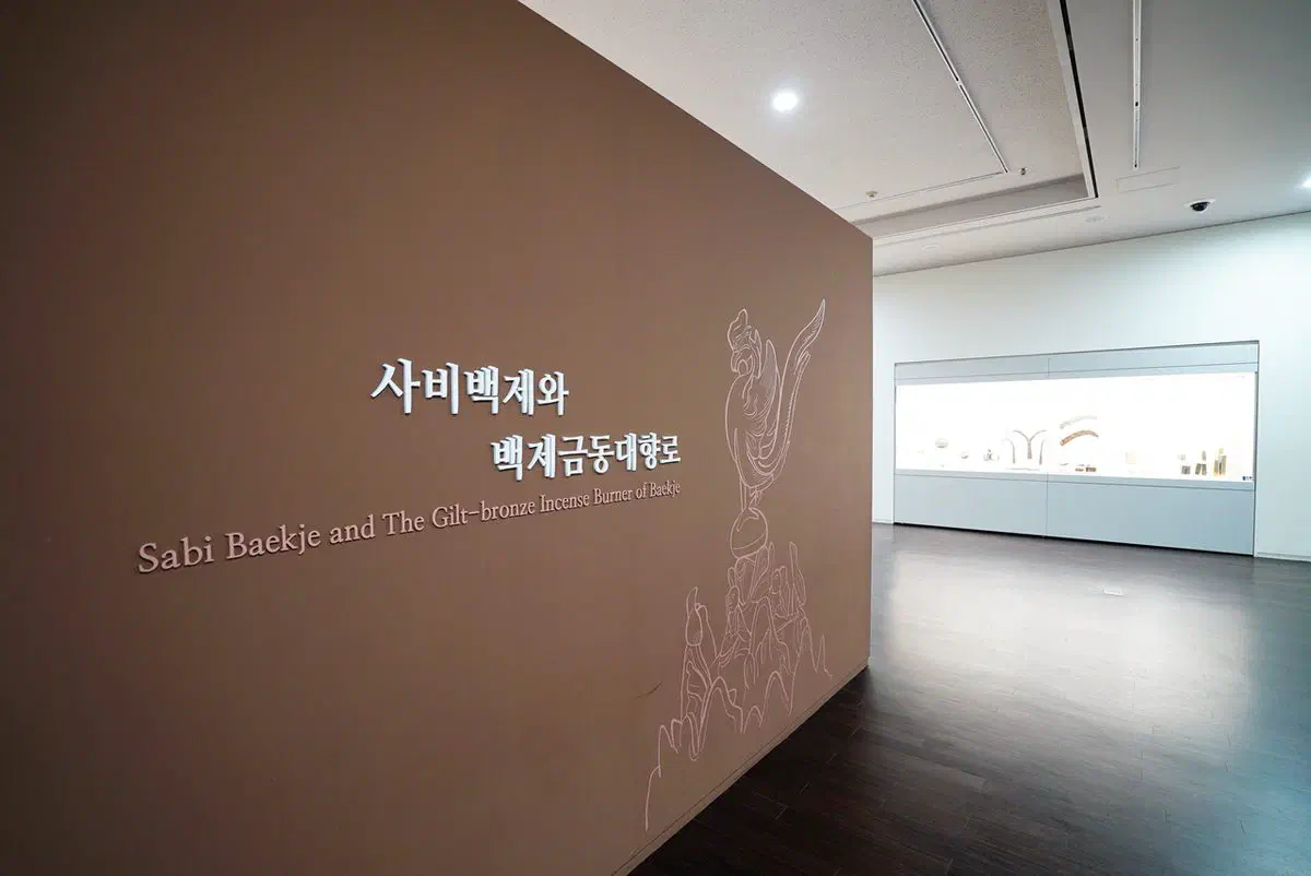 A gallery entrance wall at Buyeo National Museum headed Sabi Baekje and The Gilt-bronze Incense Burner of Baekje, with a line drawing of the censer — a phoenix above a mountain landscape — etched across the wall and a lit display case beyond