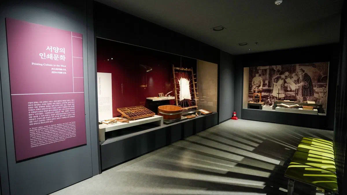 The Printing Culture in the West gallery at Cheongju Early Printing Museum: a reconstructed Gutenberg-era wooden press and type cases behind glass, with a large historical engraving of a European printing workshop on the far wall