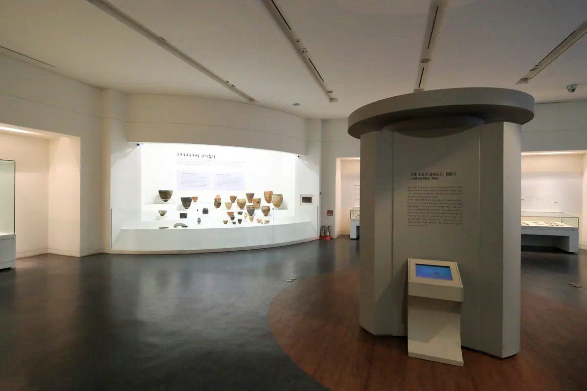 A gallery at Chuncheon National Museum: prehistoric pottery arranged along a curved lit case, with a freestanding drum-shaped interpretation column and a touchscreen stand on the polished floor