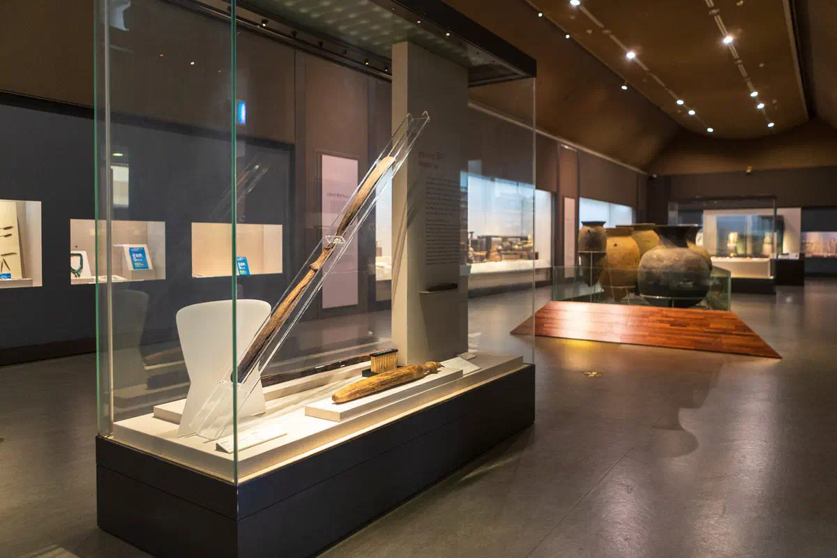 A gallery at Daegu National Museum: a lit glass case holding long slender wooden implements in the foreground, with large earthenware storage jars on a wooden plinth beyond and a warm curved ceiling above