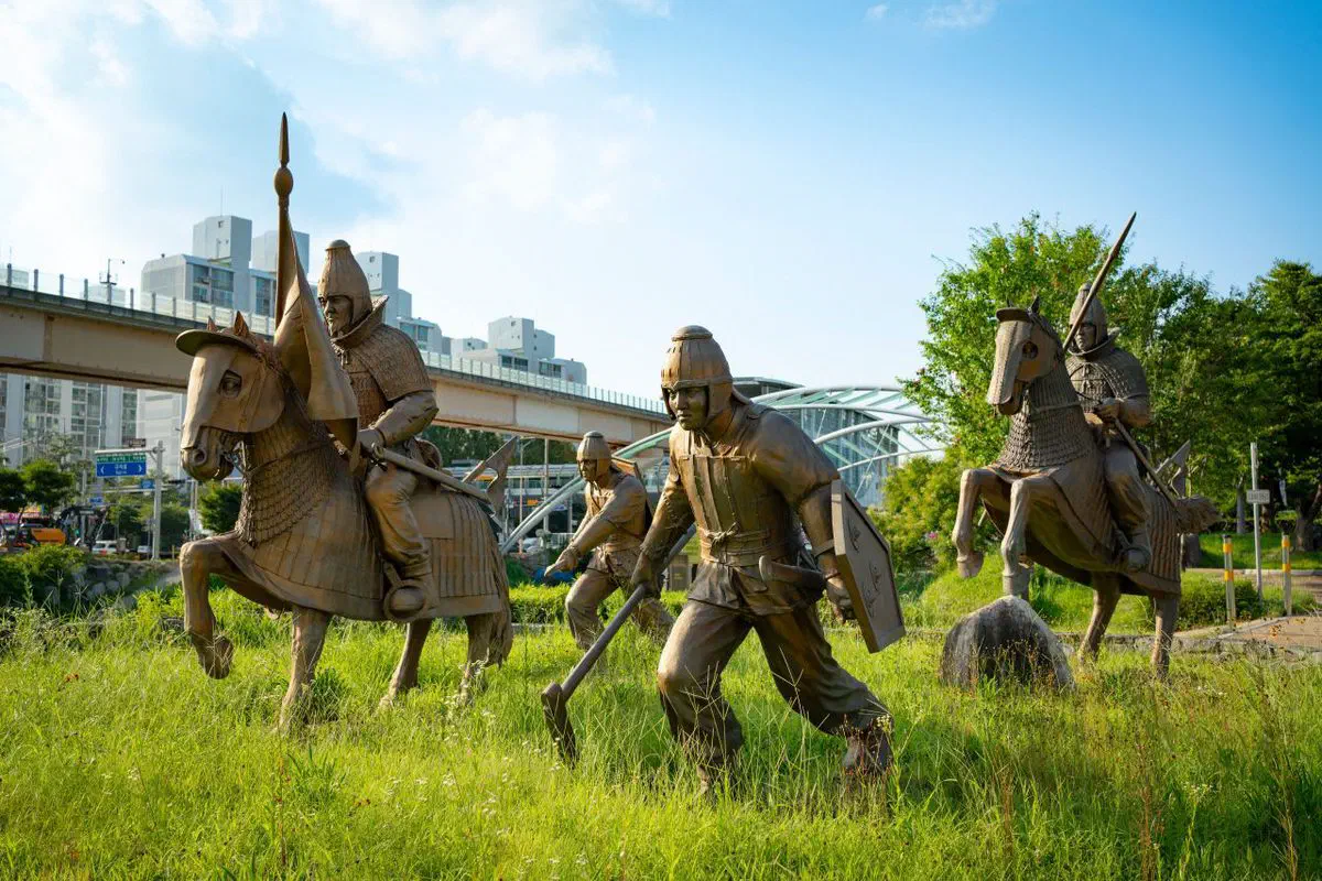 Bronze sculptures of armoured Gaya warriors and mounted cavalry standing in long grass at the Daeseong-dong site in Gimhae, with apartment towers and an elevated road behind them