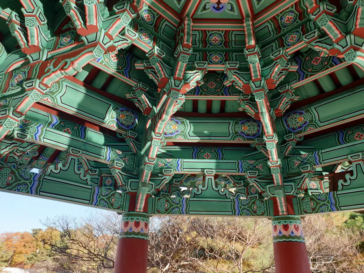 Looking up into the fully painted ceiling of a hexagonal pavilion at Deoksugung's Daehanmun gate, Seoul, with red columns and autumn trees below