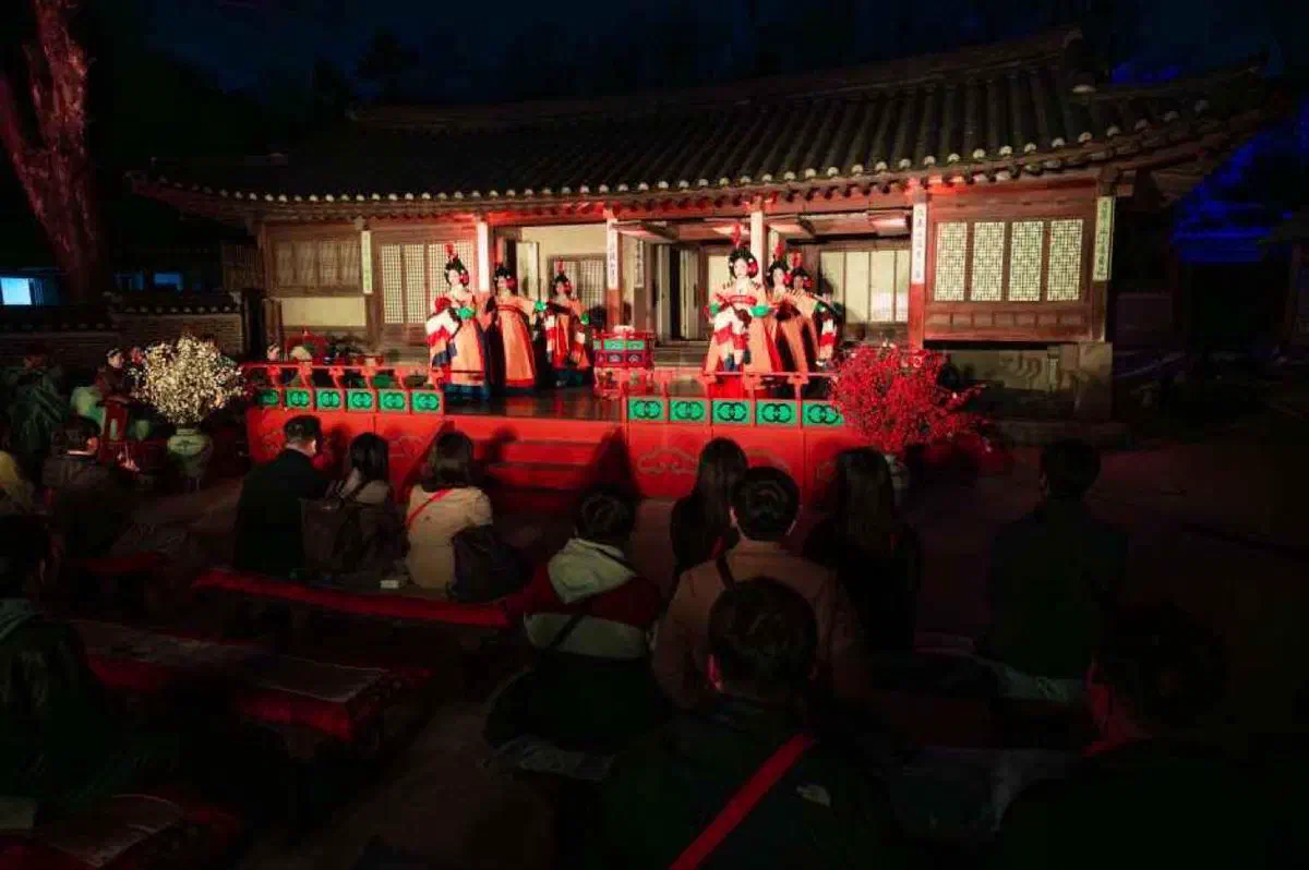 Court musicians and dancers in hanbok performing on a lit stage in front of a palace hall at night, with a seated audience watching, during Changdeokgung's Moonlight Tour