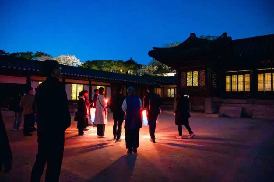 Visitors carrying lanterns across a Changdeokgung courtyard at blue hour, lit hanok windows behind them, on the Moonlight Tour