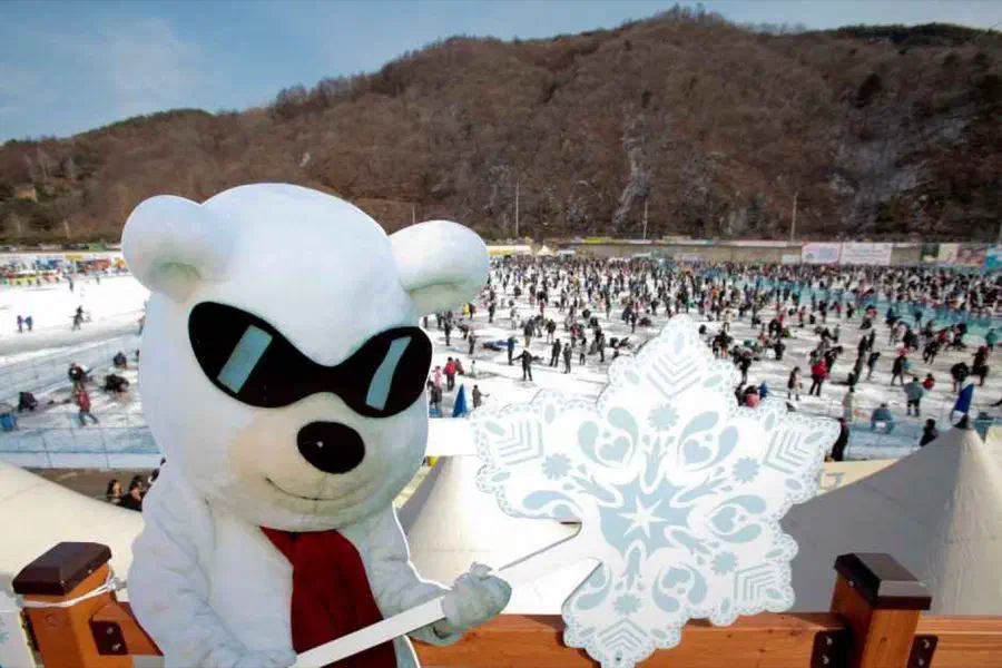 The Hwacheon Sancheoneo Ice Festival mascot in the foreground, with thousands of people spread across the frozen river behind it fishing through holes in the ice