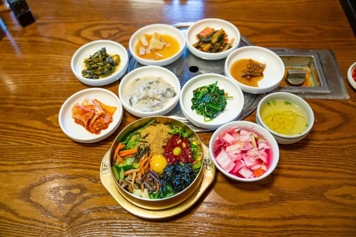 A Jeonju bibimbap set: a brass bowl of rice topped with arranged namul, gochujang, seaweed and a raw egg yolk, ringed by nine small dishes of banchan and a bowl of clear soup on a wooden table