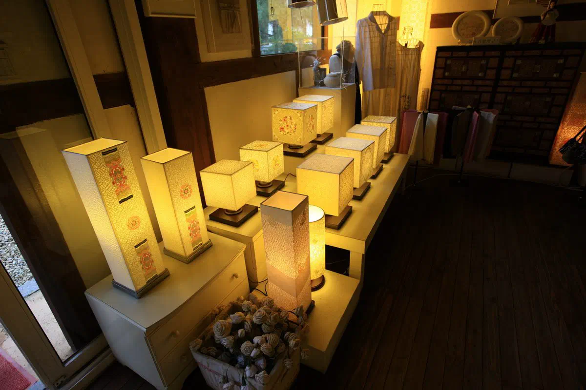 A row of lit hanji lamps in a Jeonju craft workshop showroom, their paper shades glowing warm with floral patterns, with paper roses in a basket and hanji garments on a stand behind