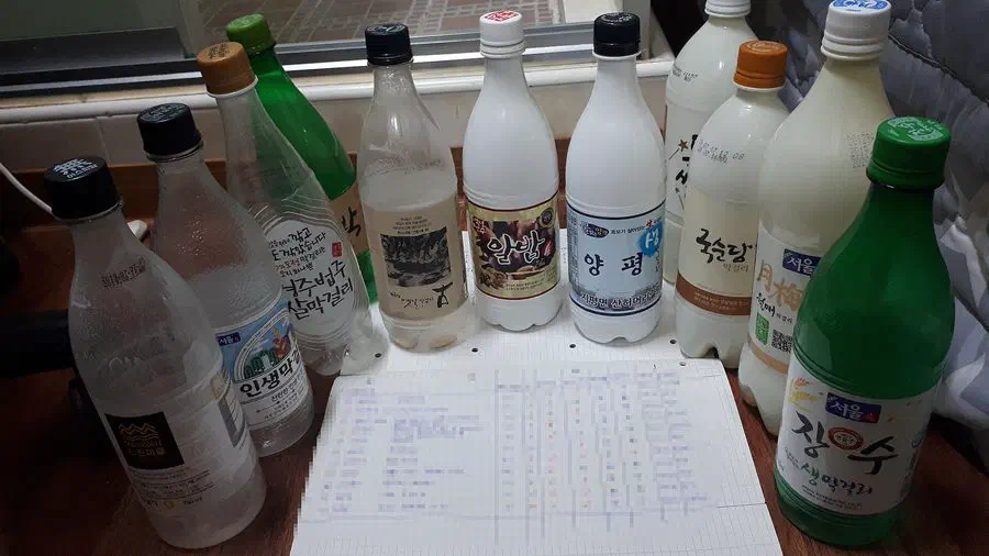 Eleven different bottles of makgeolli from separate breweries lined up on a table, most in white or clear plastic with printed labels, with a handwritten tasting sheet laid in front of them