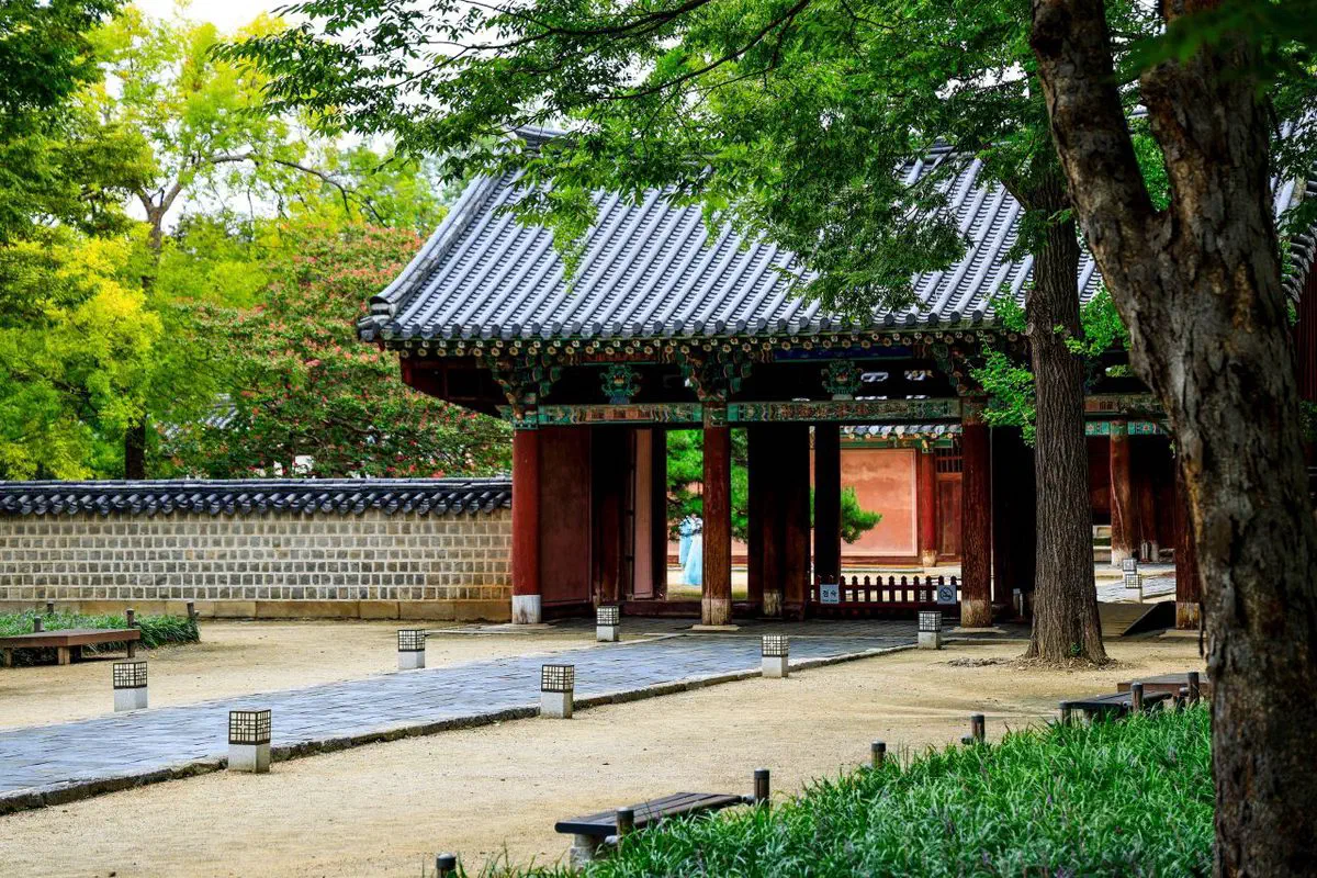 A painted gate at Gyeonggijeon in Jeonju, its beams in green and red dancheong under a tiled roof, with a tiled boundary wall running to the left and the red-walled main hall visible through the gateway