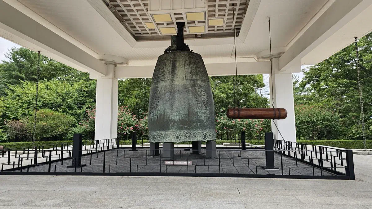 A large bronze bell on display in an open-air pavilion at the National Gyeongju Museum, with its wooden striker hanging beside it