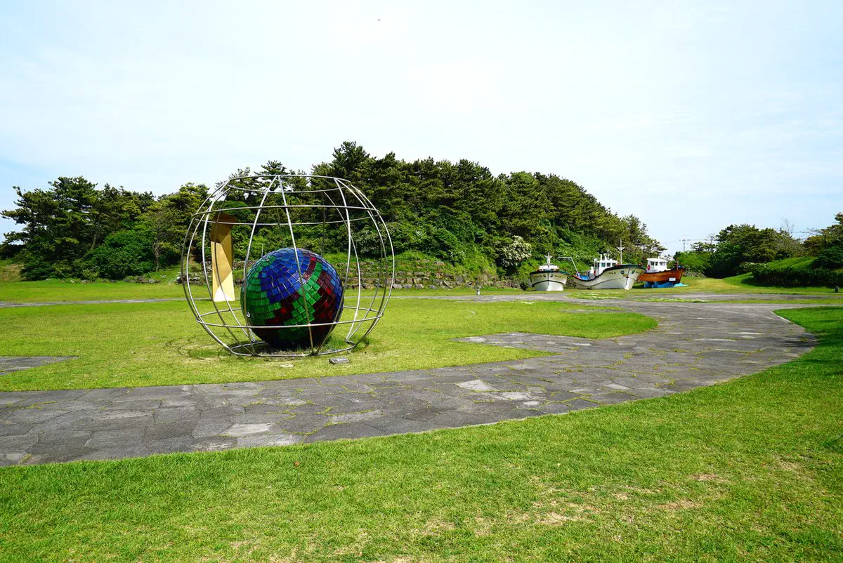 Wooden fishing boats and a mosaic globe sculpture on the museum's outdoor grounds, with Sehwa's coastline behind