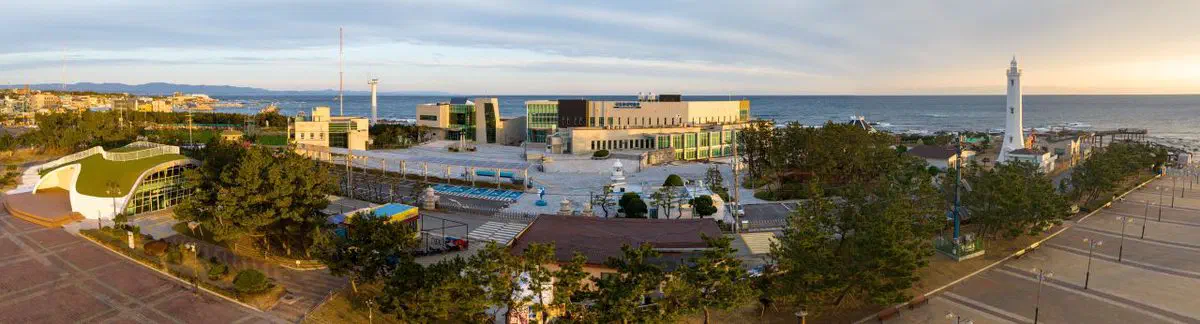 Aerial panorama of Homigot Sunrise Square at dusk, the white lighthouse tower at right and the museum's curved green-roofed hall among the buildings on the left