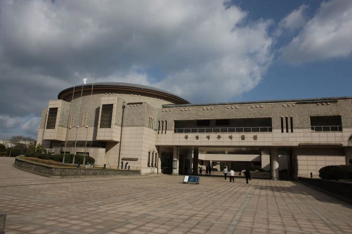 Jeju National Museum: a low pale-stone building with a shallow domed roof over one wing and textured stone cladding, seen across a wide paved forecourt with a few visitors walking toward the entrance