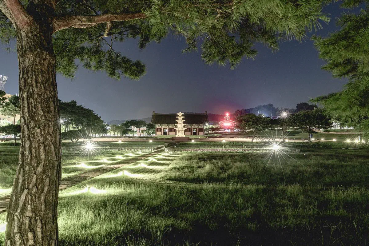 The Jeongnimsa temple site at night: the five-story stone pagoda lit in front of a single hall, with the excavated foundation stones of the vanished temple picked out by lights across the grass