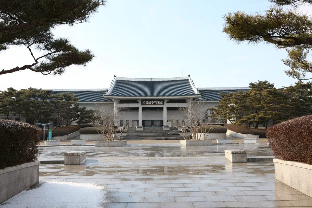Jeonju National Museum on a winter morning: a long building under a traditional tiled roof at the far end of a wide, wet, completely empty stone forecourt, with pines and patches of snow