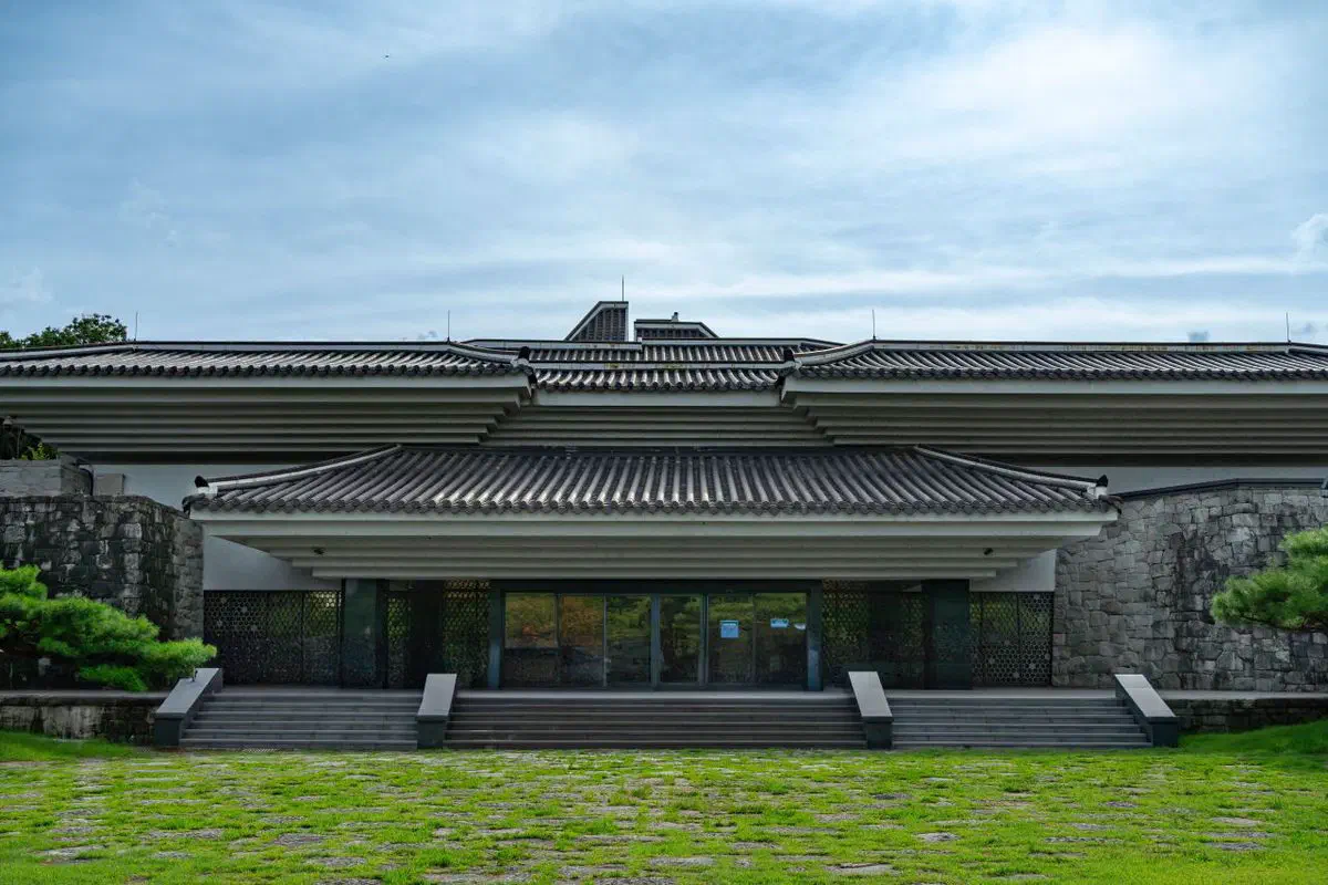 Jinju National Museum: a symmetrical building of stacked tiled roofs stepping up in tiers like a wooden pagoda rendered in stone, set into the stone rampart of Jinjuseong fortress on both sides, with a grass forecourt in front