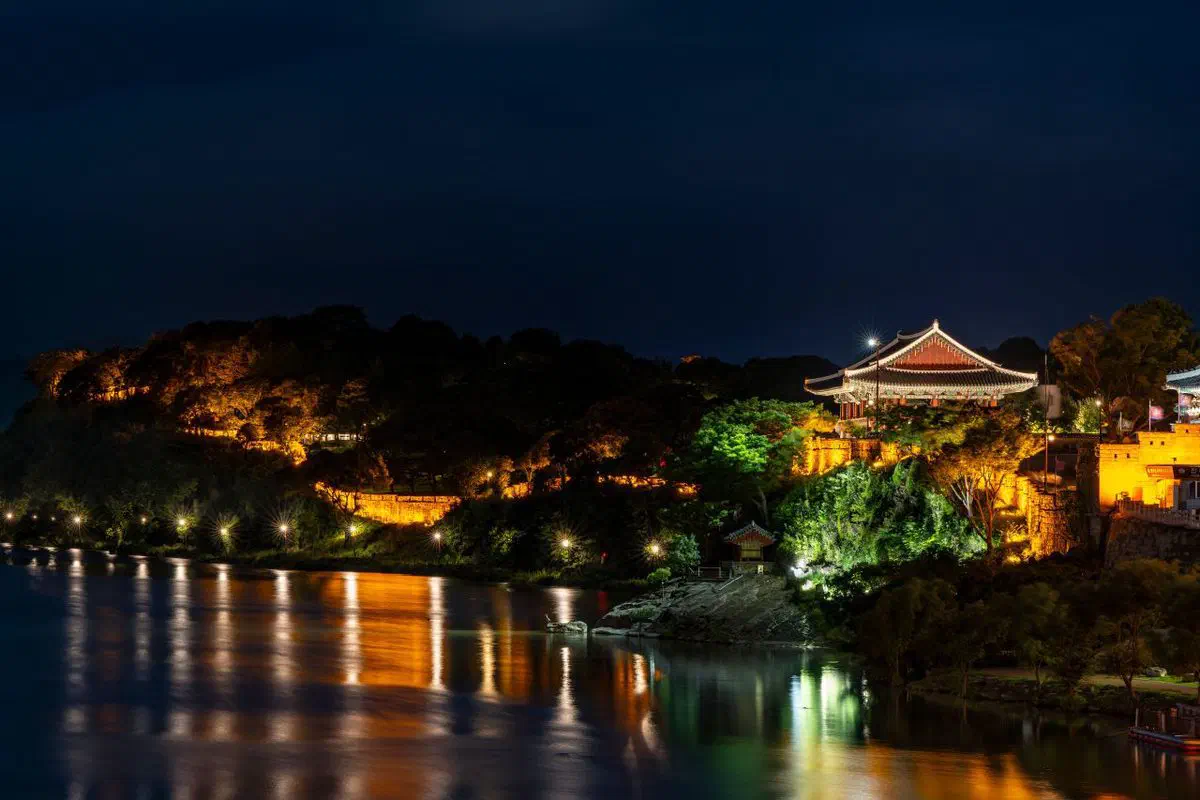 Chokseongnu pavilion lit up at night on the cliff of Jinjuseong fortress, its roof and the fortress wall glowing above the dark Nam River, with the lights reflected in the water below