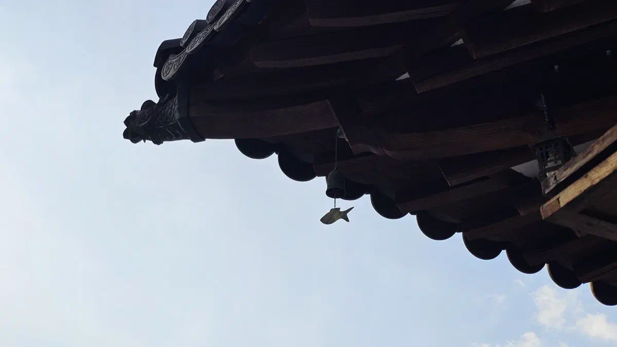 A small fish-shaped wind chime hanging from the corner of a tiled eave against the sky