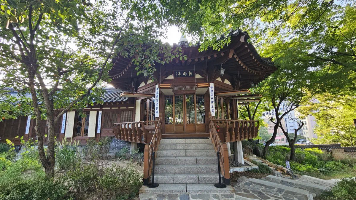 A small raised wooden pavilion among trees at Korea House, a plaque under its eaves and a stone stair up to it