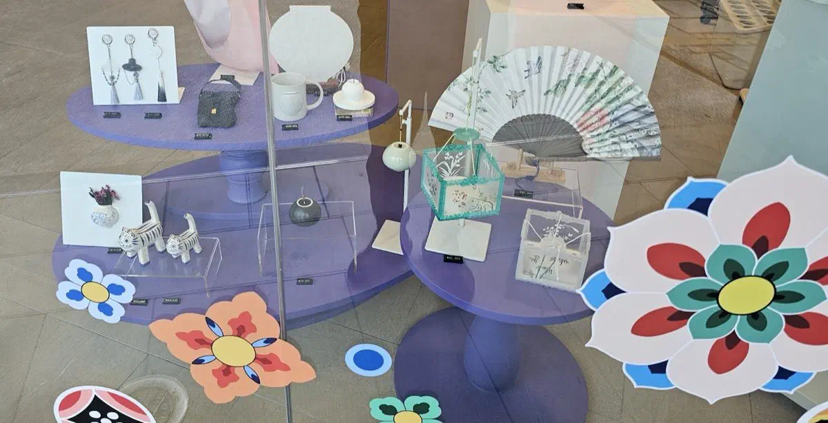 Shop window display of a fan, a white moon jar, ceramic tiger figures, tassel charms and small painted boxes on lilac stands