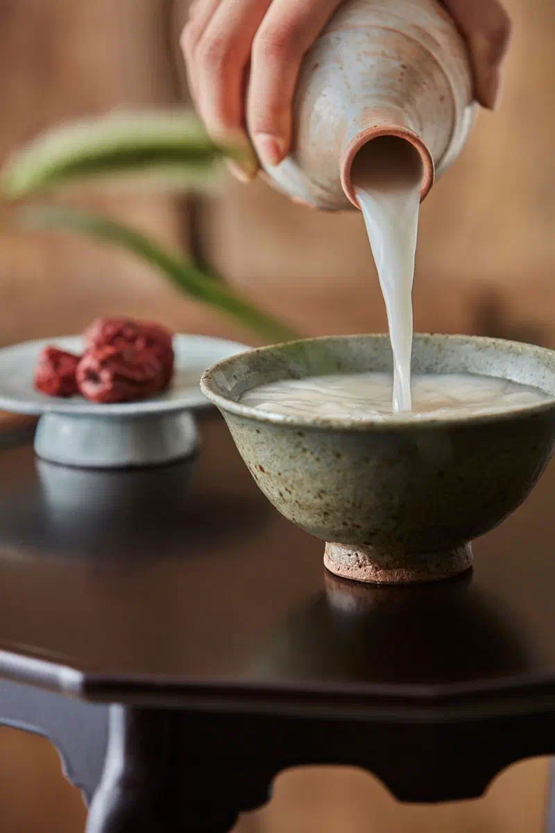 Makgeolli poured from a ceramic bottle into a shallow glazed bowl on a low wooden table, with dried jujube on a small footed plate beside it