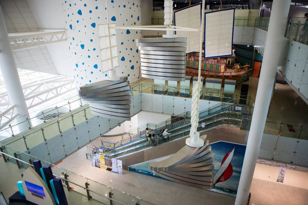 The National Maritime Museum of Korea's atrium seen from an upper floor, with a masted Joseon envoy ship displayed across the open space and escalators running between levels below it