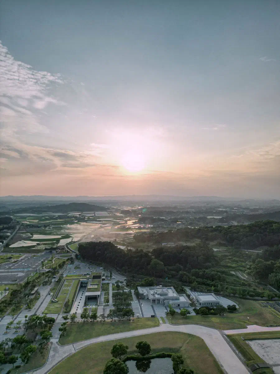 Aerial view of the Mireuksa site at sunset: the low museum building set into the ground at lower left with its roof close to grade, the open site grounds beyond it, and rice fields stretching to distant hills