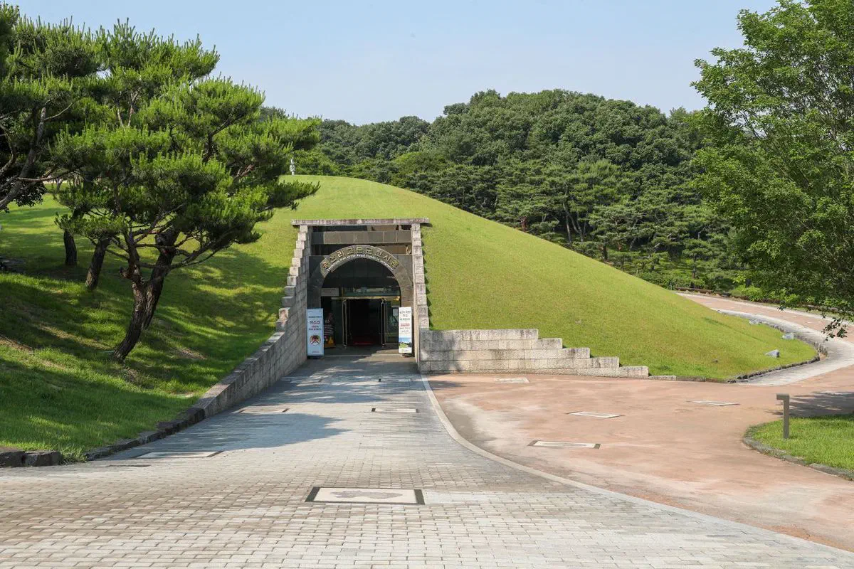The replica exhibition hall at the Gongju royal tomb park — a grass-covered mound shaped like a tomb, with an arched brick entrance set into its face and the real tomb hillside rising behind it