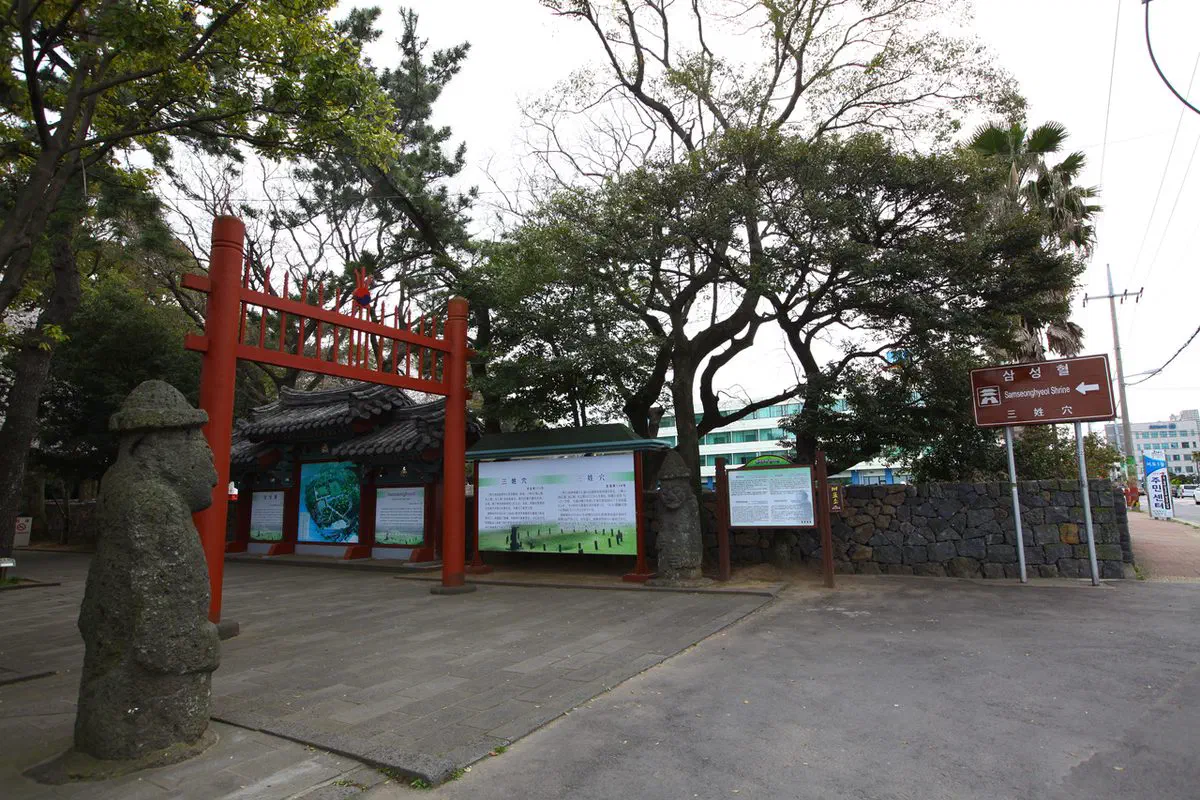 The entrance to Samseonghyeol in Jeju City: a red spiked ceremonial gate flanked by two basalt dolhareubang statues, with interpretation boards, a tiled gate building behind and a volcanic rubble wall running along an ordinary street