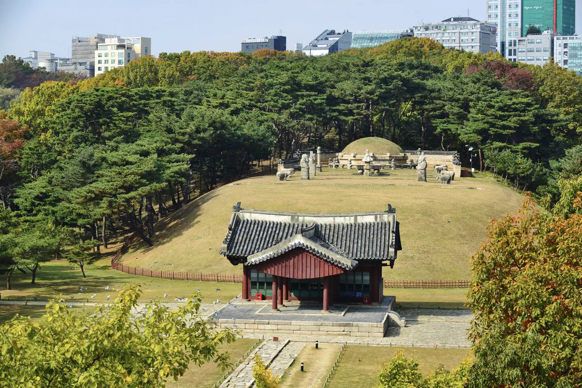 A Joseon royal burial mound, stone figures and a red shrine hall at Seolleung and Jeongneung, with Gangnam's office towers rising directly behind the tree line