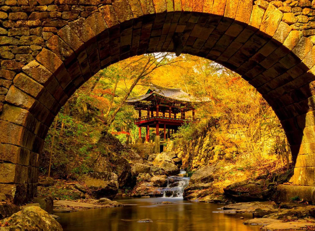 Seungseongyo, Seonamsa's arched stone rainbow bridge, framing a small wooden pavilion and a stream beyond it in autumn