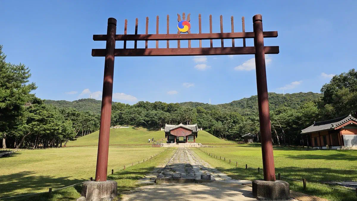 A red wooden hongsalmun gate with a cut-out ornament across the entrance path, the ritual hall and mound beyond