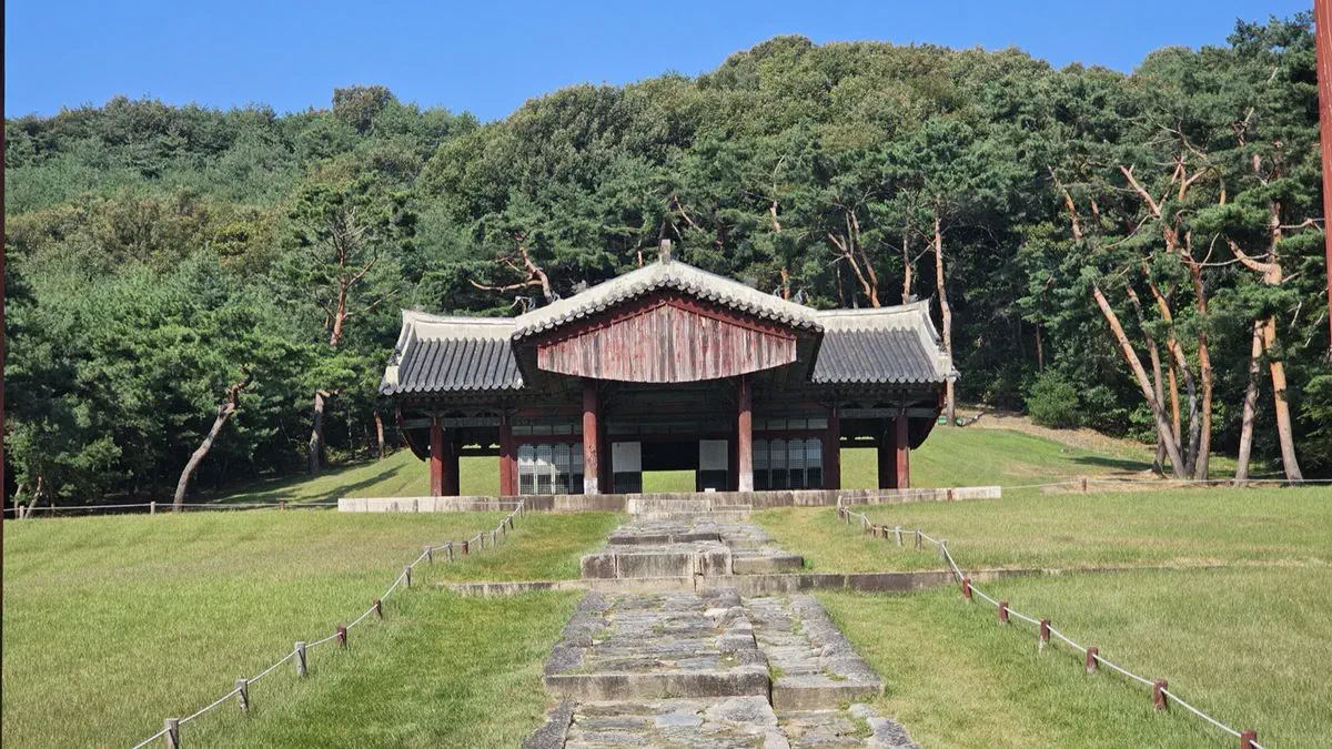 The T-shaped ritual hall of a royal tomb at Seooreung at the end of a stone path, rope fences along the grass and a pine-covered hill behind
