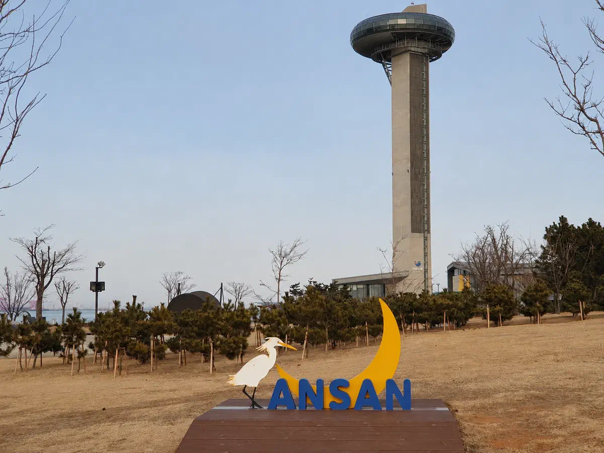 The 75-meter Sihwa Narae Moon Observatory tower rising from the tidal power plant on Daebudo, seen from the park below with an Ansan sign in the foreground