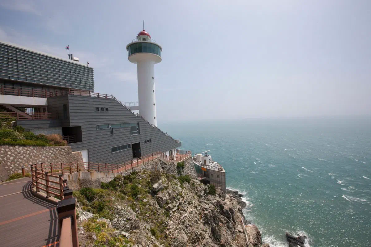 The lighthouse at Taejongdae on Yeongdo, white tower and observation building set on a rocky cliff above open sea, with a boardwalk path running along the headland